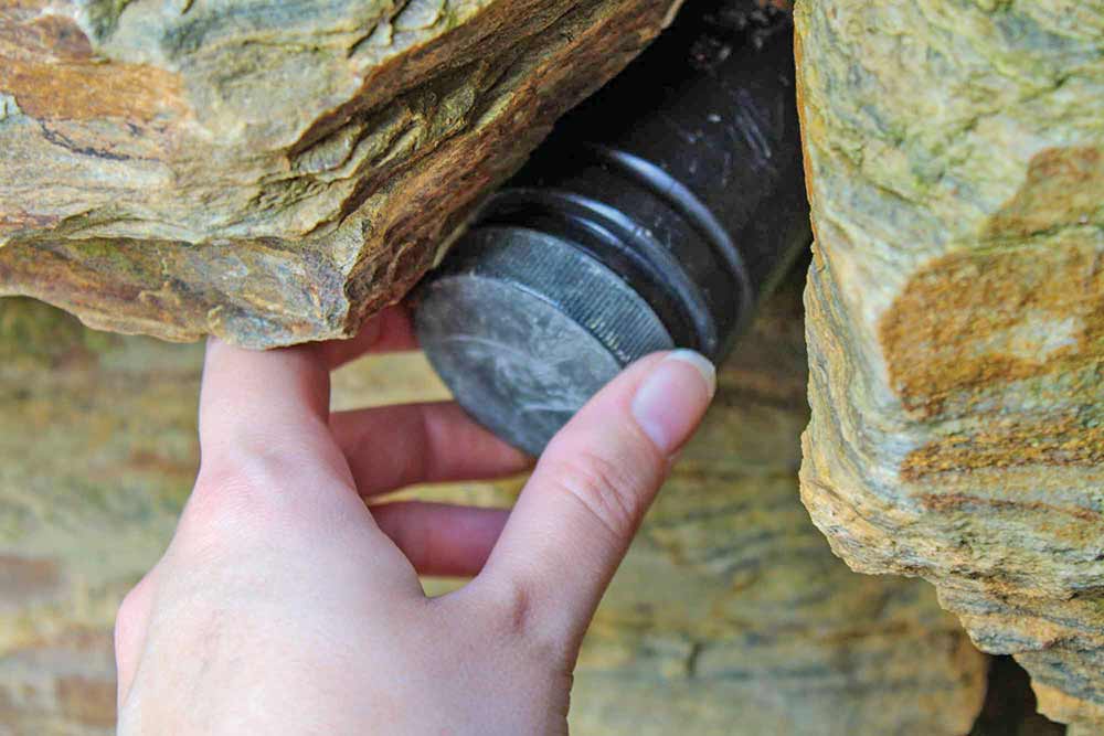 A hand reaches out to grab a black canister from inside a pile of rocks.