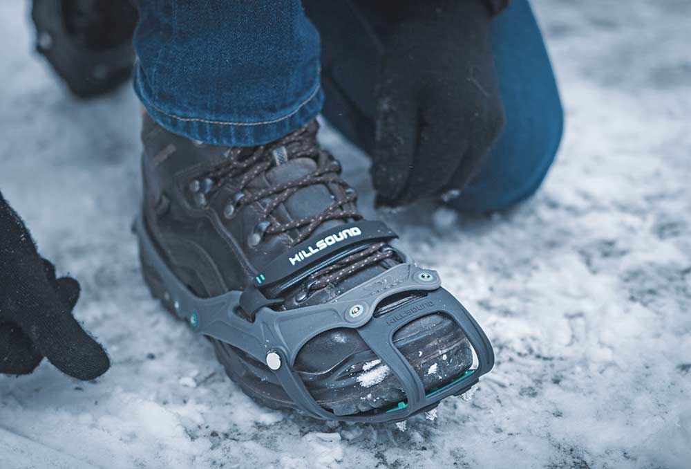 A person wearing jeans and boots straps gray rubber crampons to their boots.