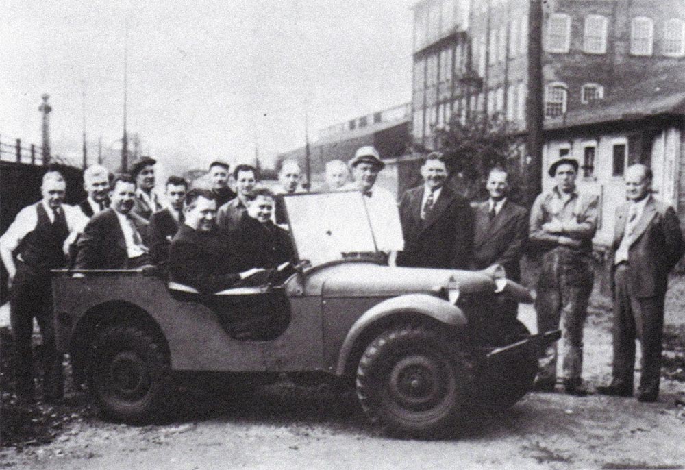 A gathering of men who helped Karl Probst design the Jeep prototypes pose behind an early Jeep while two more sit in the vehicle.