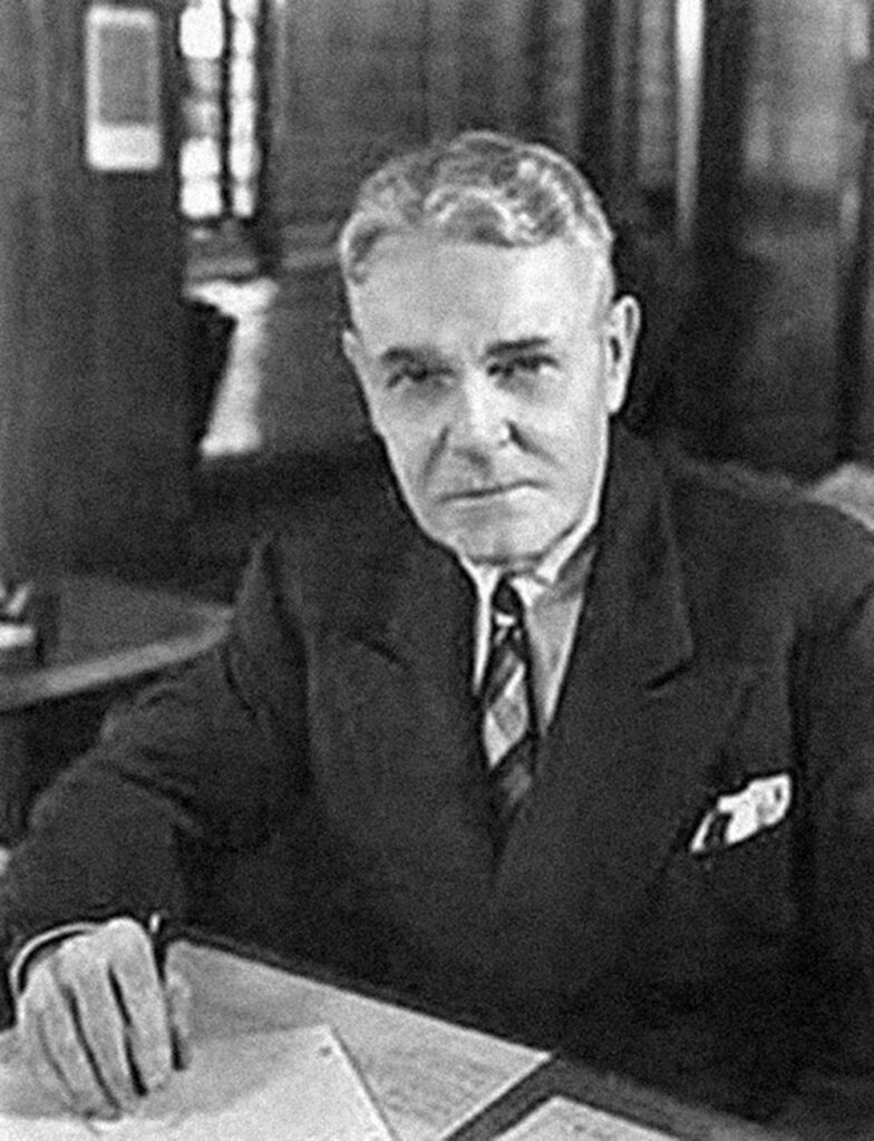 Black and white photo of Karl Probst, a middle aged white man, sitting at a desk in a suit.