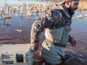 A man in camp wades through waist-deep water pulling a box behind him.