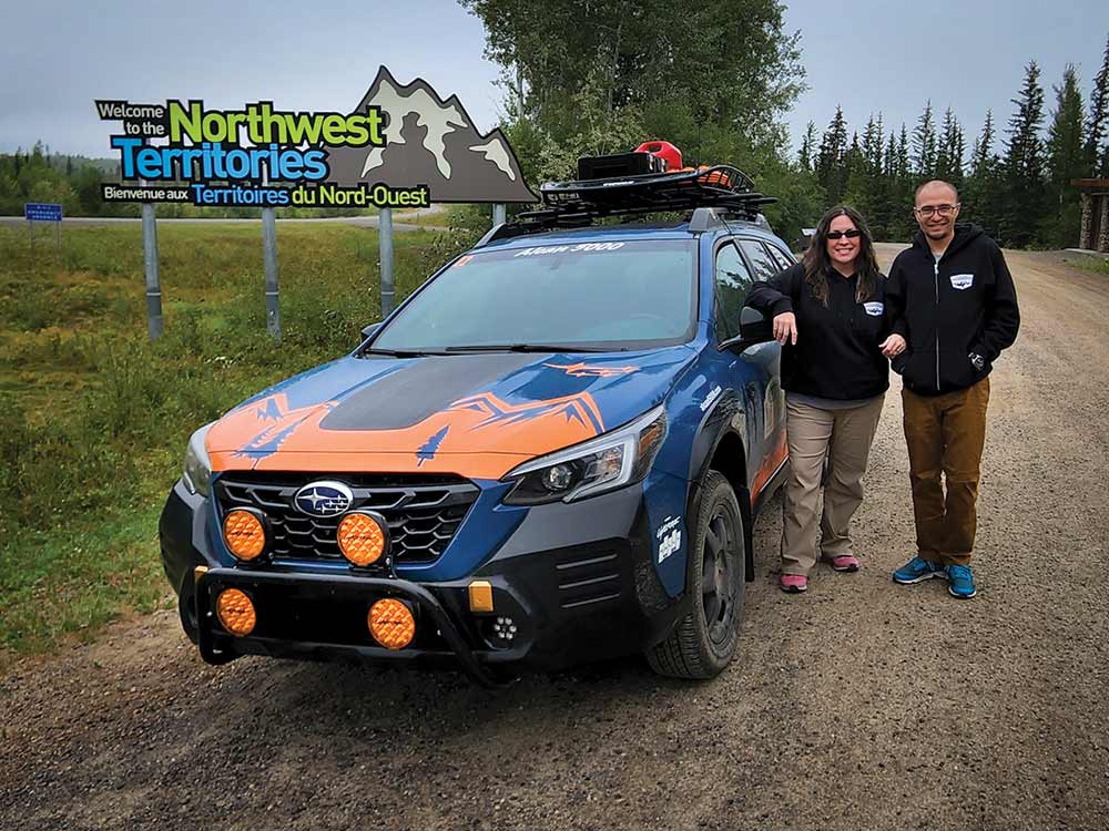 The author and her husband pose next to their Alcan Rally car, a Subaru Outback Wilderness in front of a Northwest Territories welcome sign.