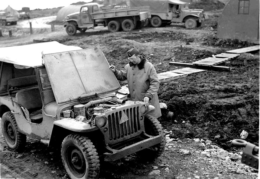 Black and white photo of a man in a coat inspecting under the hood of a doorless Karl Probst early Jeep prototype.