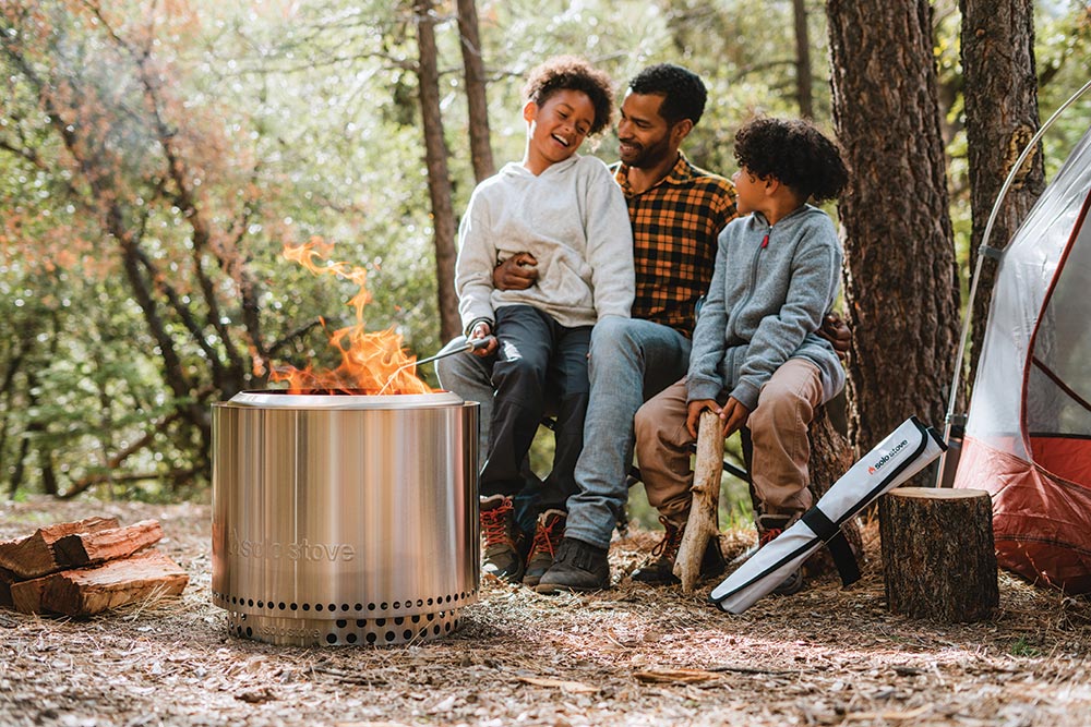 A man with two children sitting withi him roast marshmallows on sticks using the flame from the cylindrical metal fire pit.