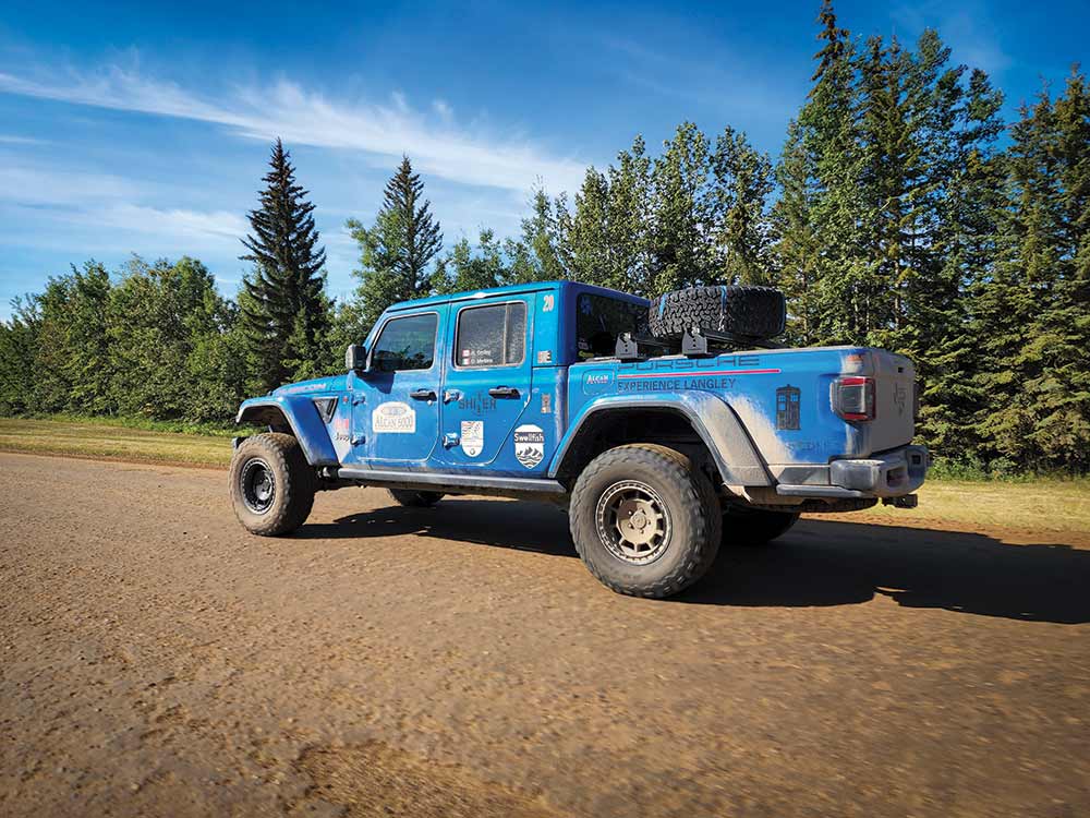 A bright blue Jeep Gladiator parks on the side of a wide, wooded dirt trail during the Alcan rally.