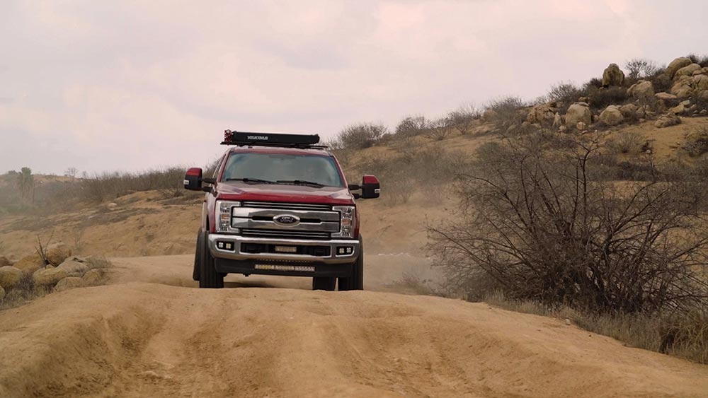 The red Ford F250 approaches from the front along a dirt path under cloudy skies.