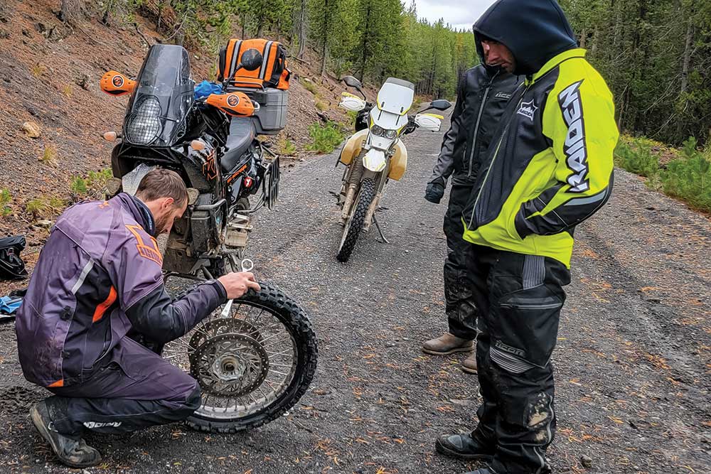 Two male bikers gather around a bike as a third man squats to fix a wheel.