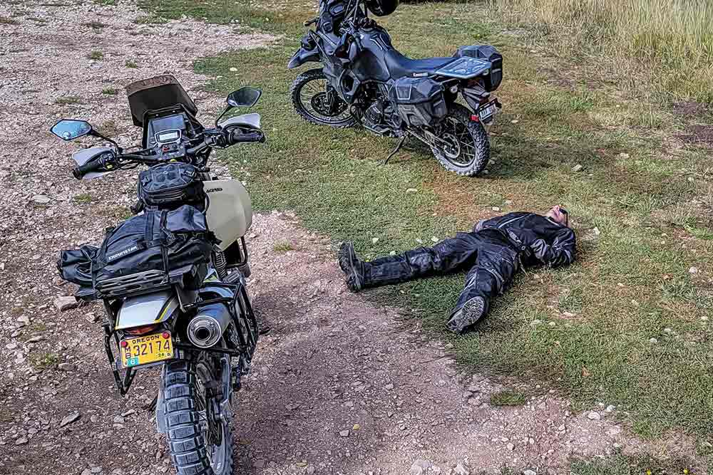 Two motorcycles park next to the edge of the trail while a man in leathers lays down on the grass to rest.