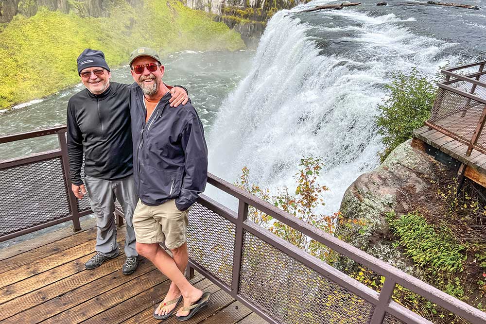 Two older men pose arm in arm at a waterfall adventure overlook. 