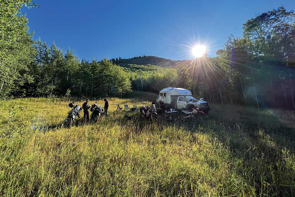 In a tall grass field under clear sky, a group of moto adventurers take a pause next to the adventure support truck.