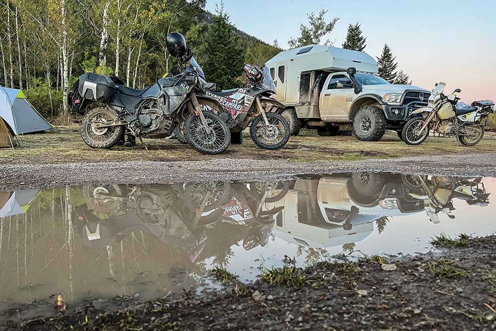 A lineup of motorcycles and the support truck with a bed camper are reflected in a puddle.