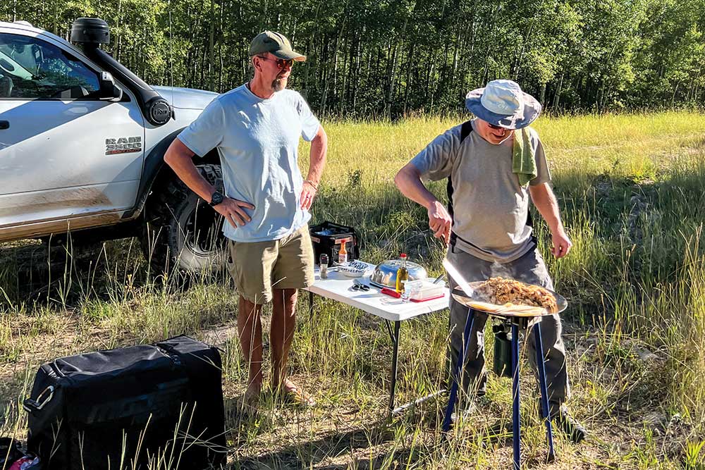 Two men stand in a grass field near the support truck cooking dinner on the adventure.