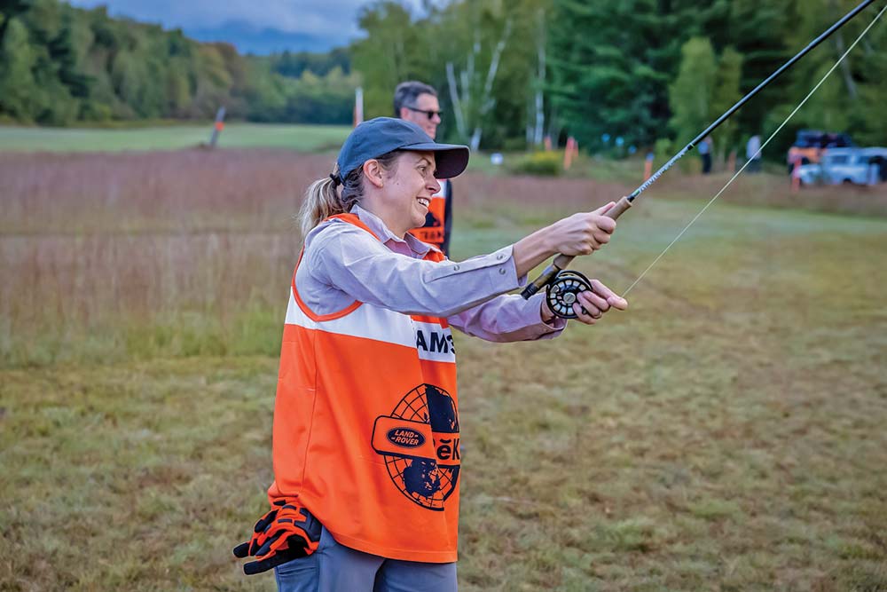 A woman in a blue ballcap and orange pinny practices her fly fishing skills.