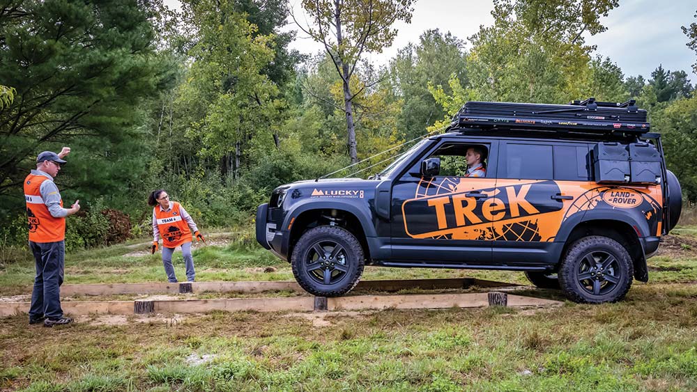 Two team members coach a Land Rover driver across a wooden balance beam.