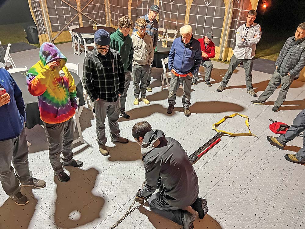 A group of Land Rover TReK competitors in hoodies watches a demonstration of how to use a Hi-Lift jack.