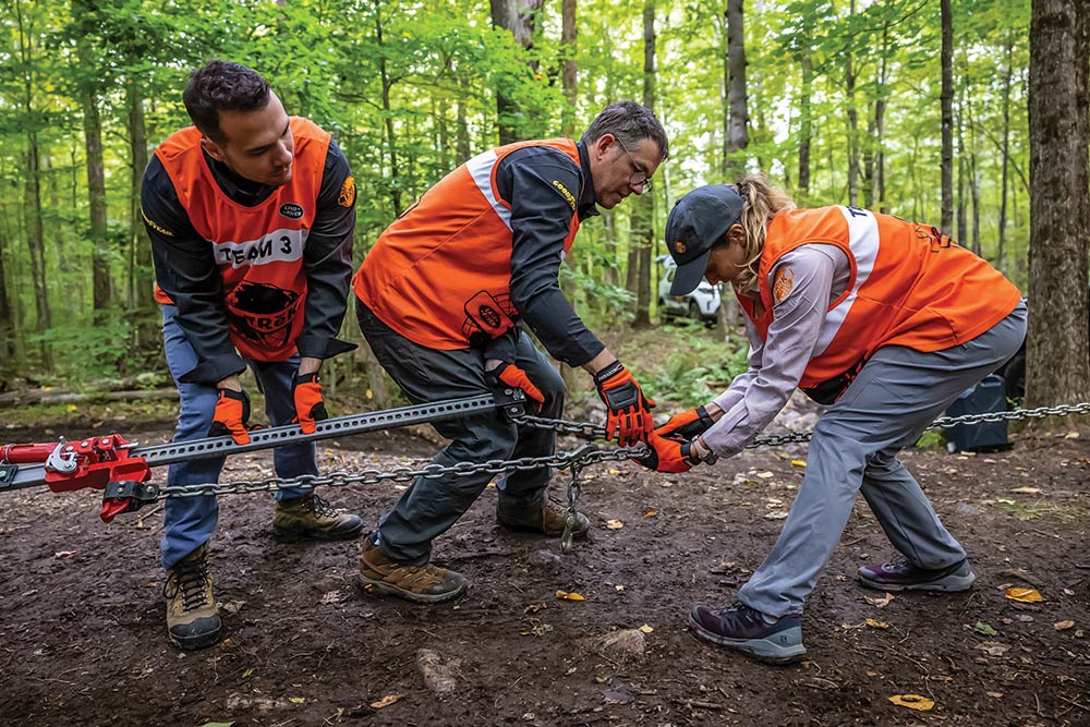 Three competitors in orange vests work together to attach chains to a hi-lift jack.