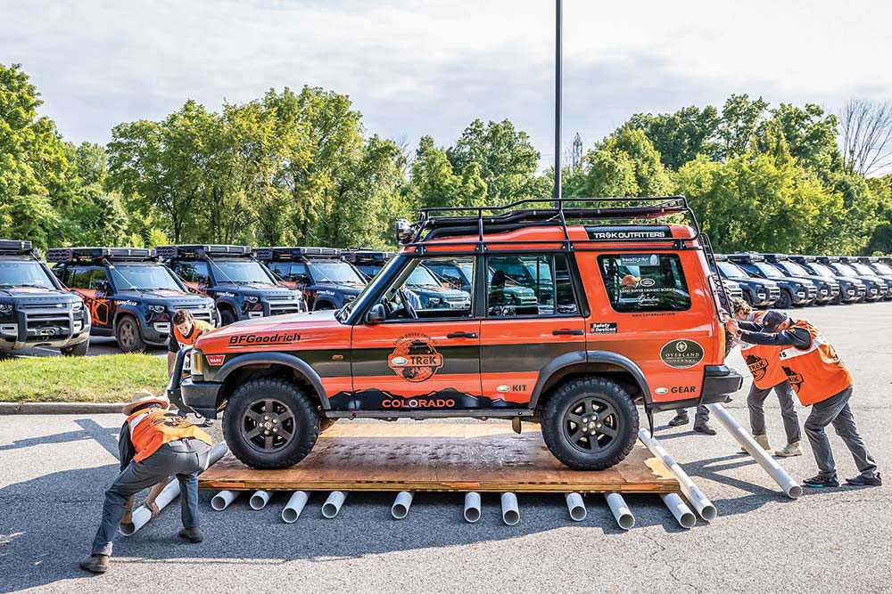 A team of competitors rolls a black and orange Defender across the parking lot on plywood and PVC pipes.