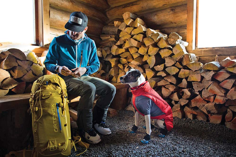 A man in a blue jacket, green gear backpack, and dog in red jacket sit in a shed with wood stacked to the side.