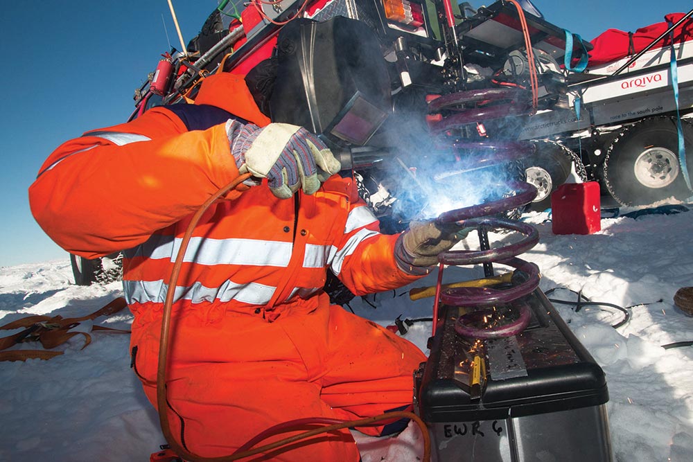 A person in a bright orange reflective snow suit and welding hood attempts to fix a broken spring.