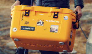 A man carries the orange Dometic cooler with lots of stickers on it.