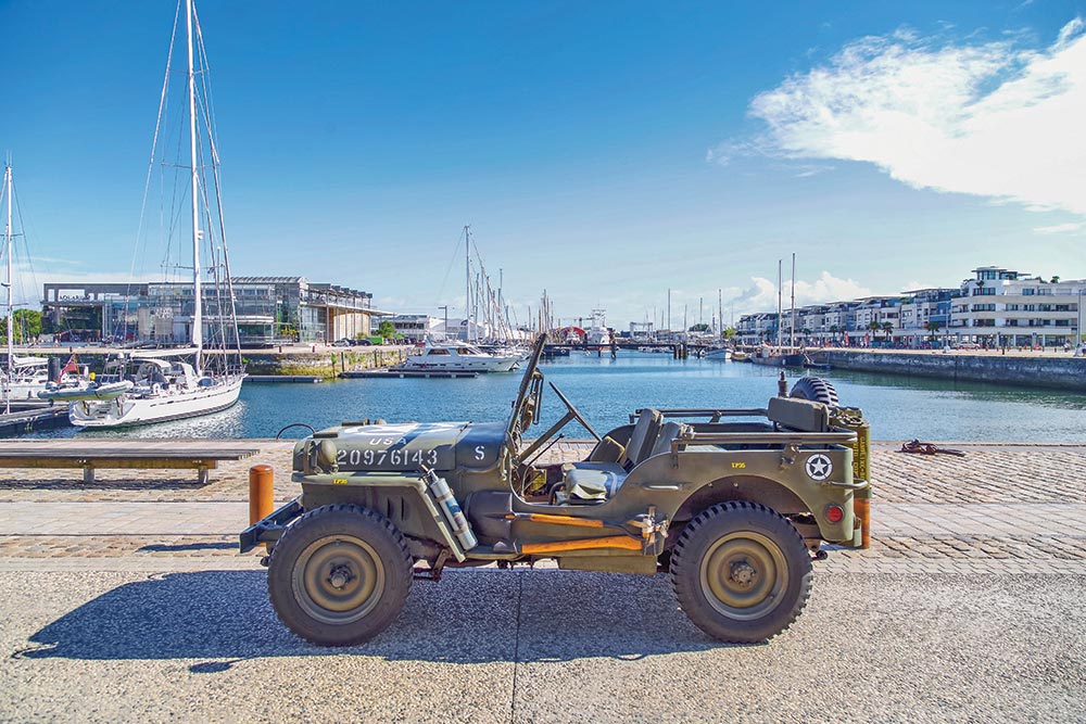 Color photo of an old military Jeep parked in front of a harbor.