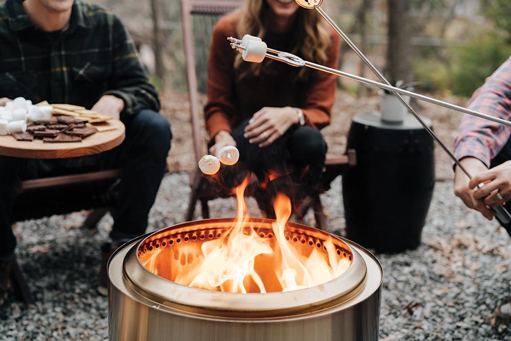 Several adults roast marshamllows on skewers around the cylindrical metal fire pit.