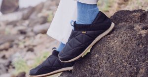 A guy in white pants and blue socks and shoes rests his feet on a black rock.