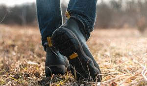 A guy in jeans and rubber boots walks through a field.