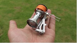 A hand holds the small sliver and orange camp stove burner.