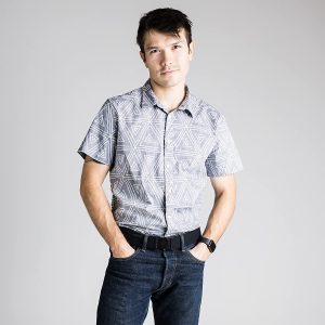 A guy in a blue button up shirt, jeans, and a black belt models for the camera.