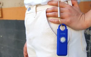 A gal in white pants holds a rectangular blue alarm on a brass carabiner.