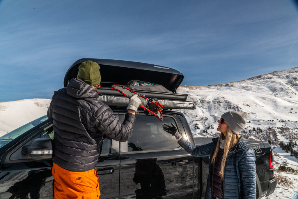 Man loads cargo box on top of truck with snow gear while woman watches.