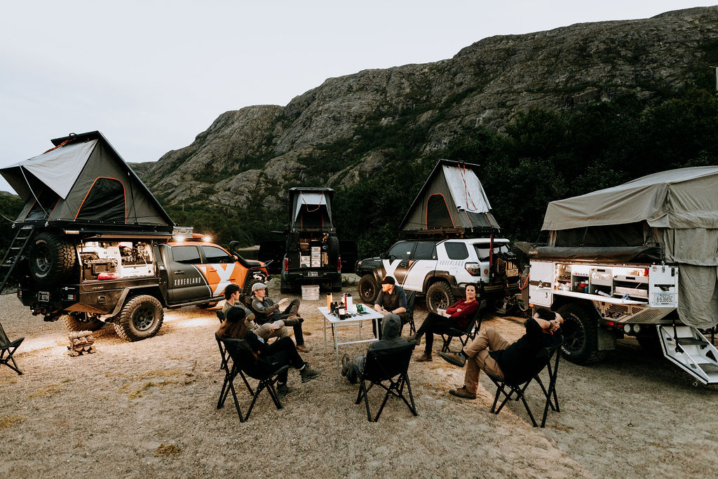 Still from xOverland's new Scandinavian series; 3 trucks with roof top tents surround a group of people seated in camp chairs around a folding table.