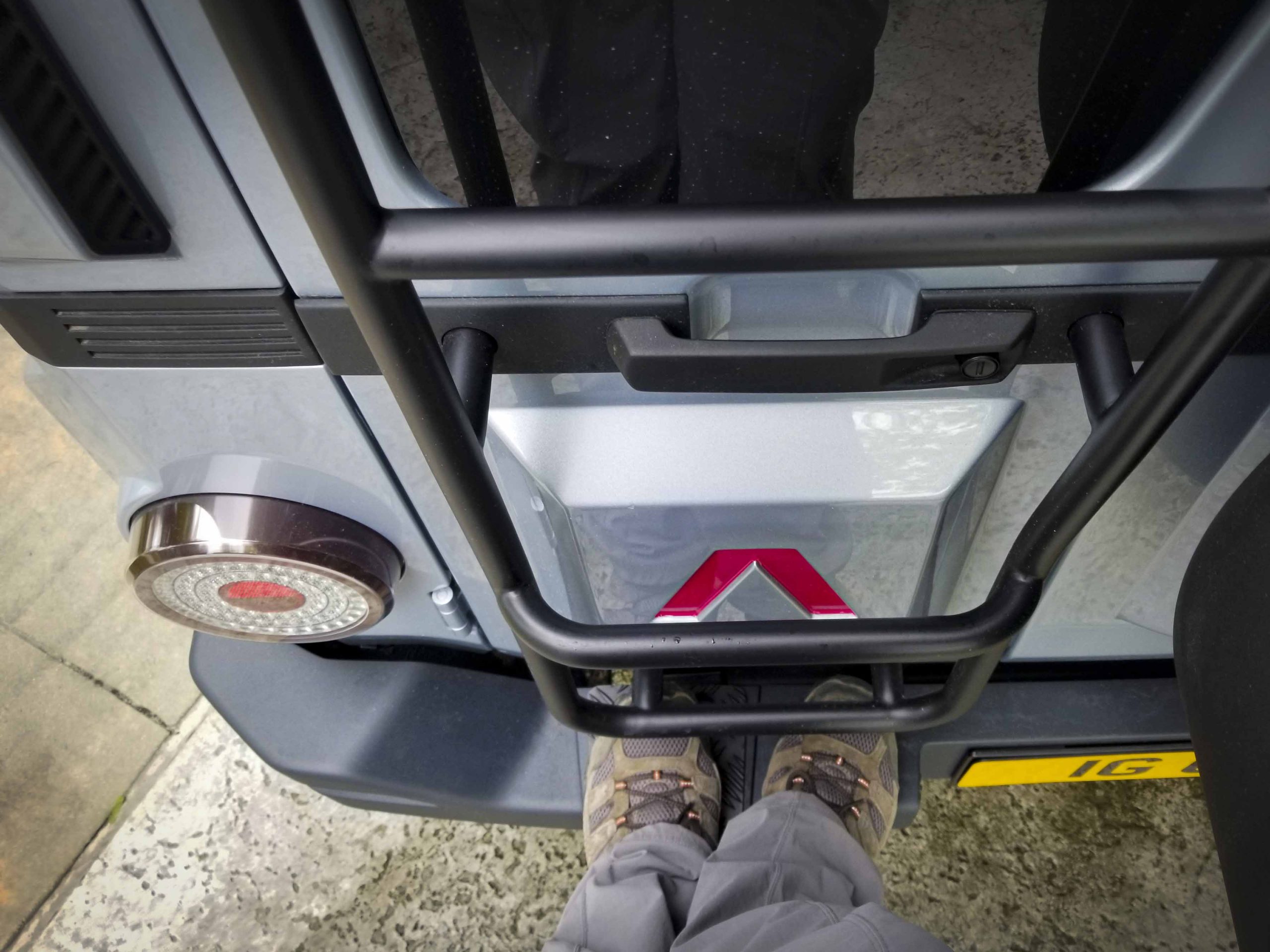 Feet stand on the bumper below the ladder on the rear end of an INEOS Grenadier.