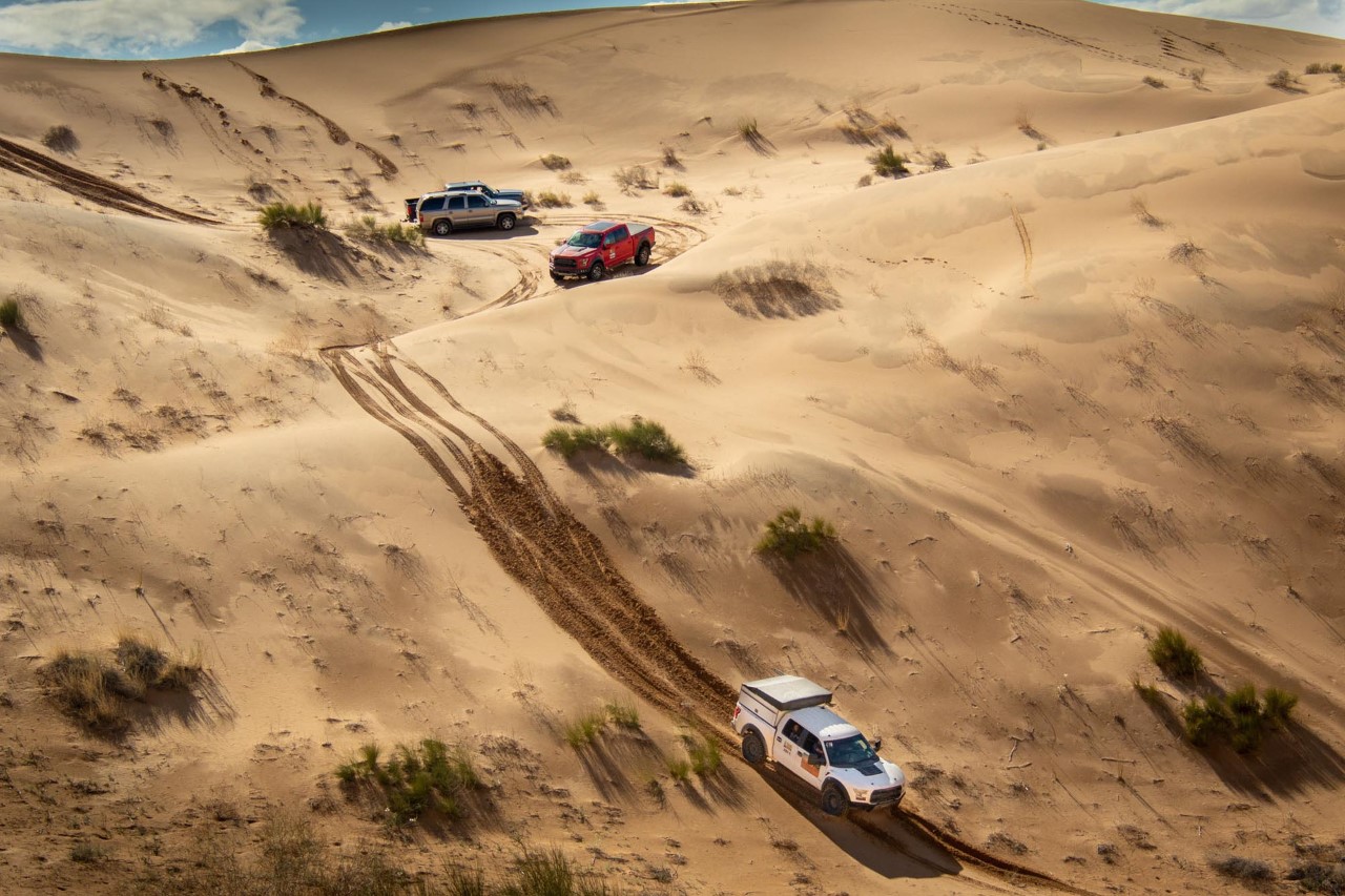 Three of last year's Adventure Raid participants in trucks wind down a sandy hill in the desert.