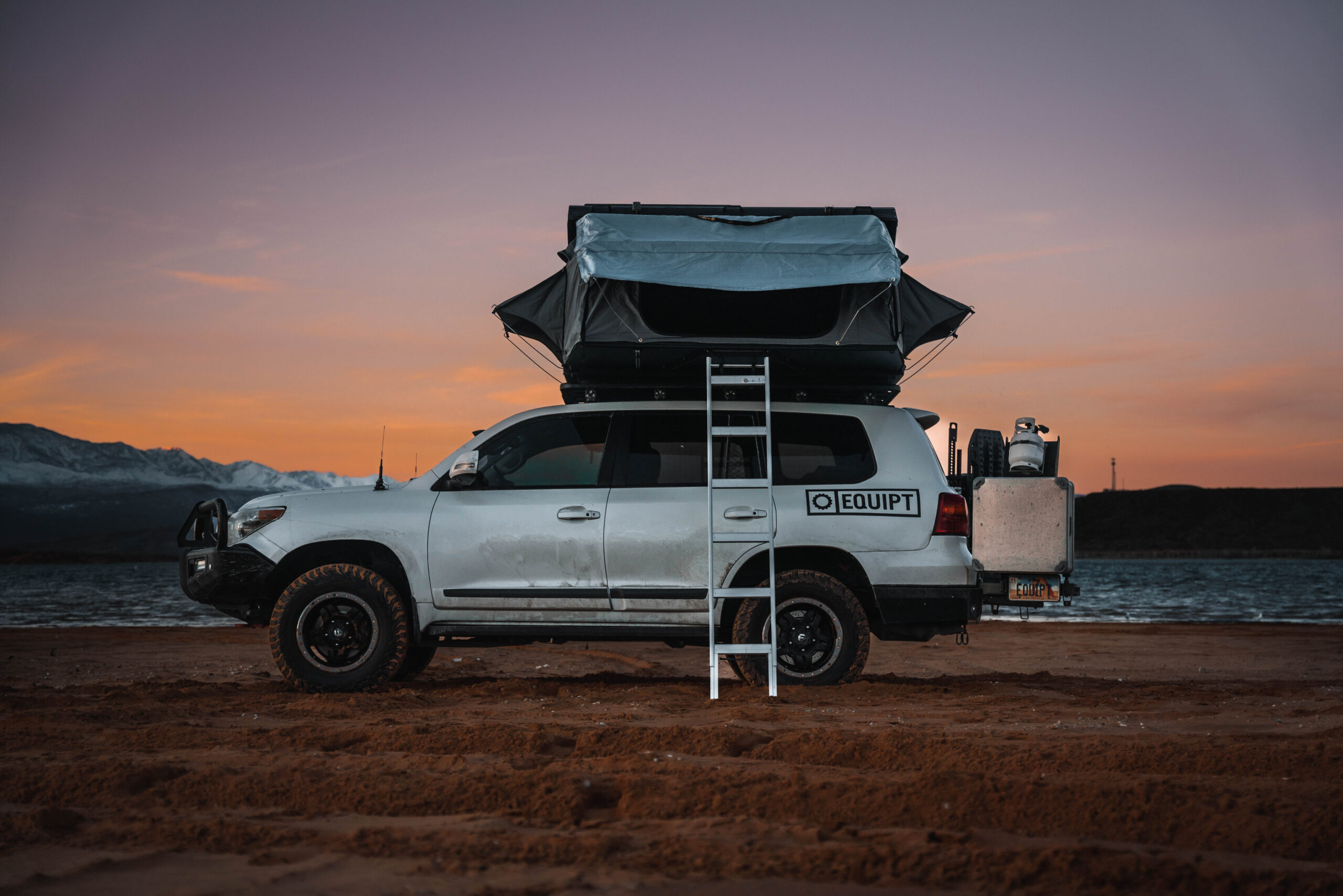 An SUV parks on dirt in front of a pink sunset with the Eezi-Awn roof top tent deployed.