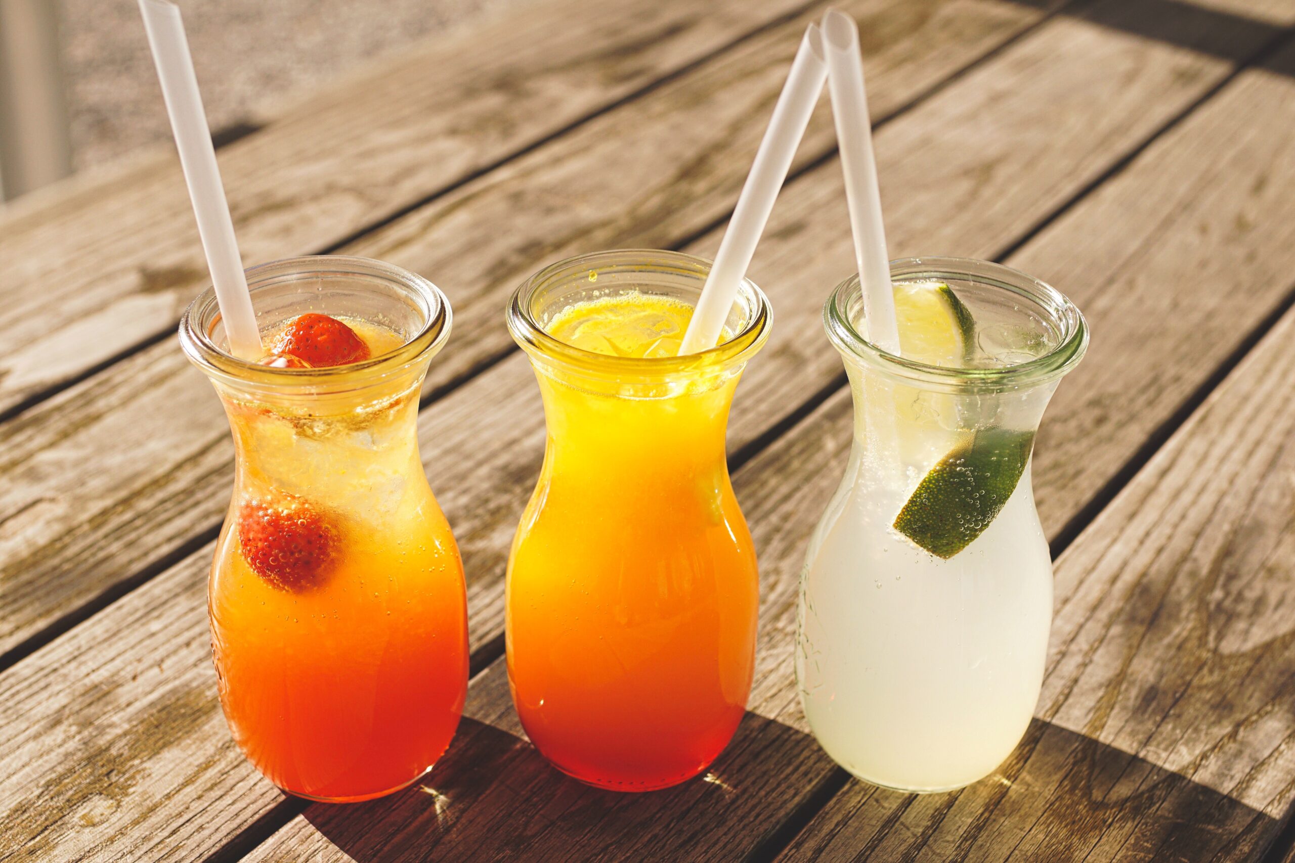Three cold summer drinks with straws sit on a wood table. Two are orange and red in color, and one is white.